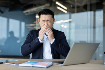 Sick young Asian man sits at the desk in front of a laptop in the office in a business suit and...
