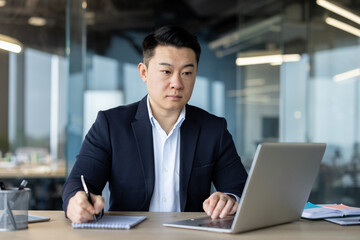 A serious and focused young Asian man in a business suit is sitting in the office at the desk, writing in a notebook and looking at the laptop screen