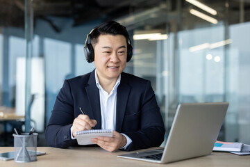 A young Asian male businessman in a suit and headphones is sitting in an office at a desk, looking at a laptop screen and making notes in a notebook