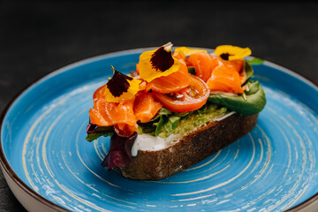 Gourmet smoked salmon toast with fresh vegetables and edible flowers on a vibrant blue plate