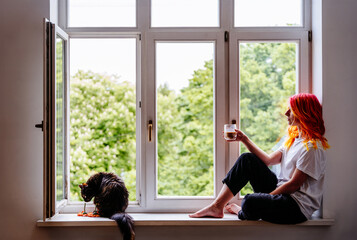 Woman enjoying beverage by window with cat on sunny day