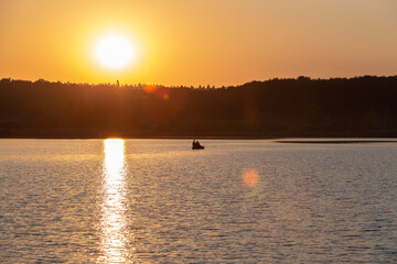 A serene sunset over a tranquil lake with two people boating in the distance, surrounded by trees and peaceful water reflections