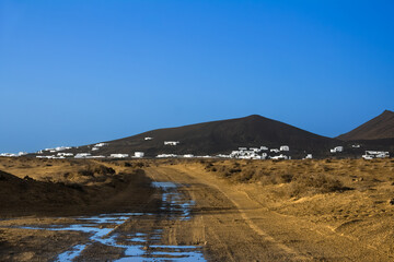 Puddles dot the road leading through a desert landscape toward a town of white traditional houses perched on a volcanic slope in Lanzarote, Canary Islands, Spain.