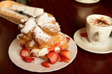 Choux pastry with whipped cream and strawberries on a plate, with a cup of cappuccino nearby.