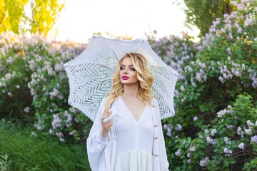 Young beautiful woman in white historical dress and headpiece standing with umbrella among trees with lilac flowers.