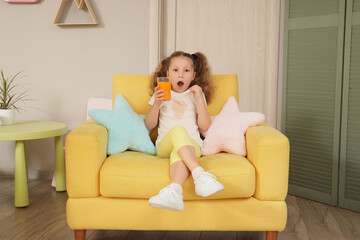Obraz premium Shocked little girl spilled juice on her t-shirt in armchair at home