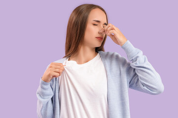 Displeased young woman in dirty clothes covering her nose on lilac background, closeup