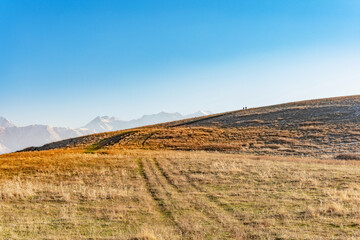 Small dark silhouettes of two people and a dog coming down a hill against the background of high mountains and a clear blue sky. There is yellow dry autumn grass on the hill.