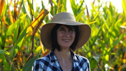 Happy smiling female farmer looks into camera standing near corn field. Portrait of adult beautiful agronomist in straw hat with maize meadow at background. Agricultural business concept. Close up