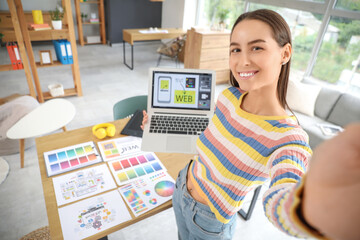 Young female graphic designer with laptop taking selfie in office, closeup