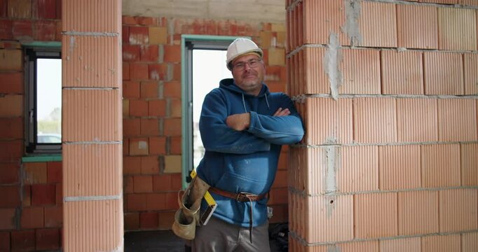 Smiling construction worker in blue hoodie, arms folded, leaning against brick wall in unfinished building, safety helmet on, industrial site background