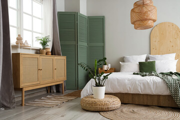 Interior of light bedroom with cozy bed, houseplants and green folding screen
