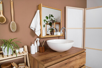 Modern sink, chest of drawers, folding screen near brown wall in interior of bathroom