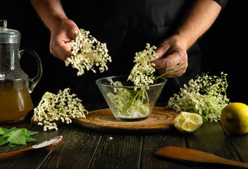 In a cozy kitchen, hands delicately place fragrant elderflower blooms into a glass bowl. A wooden...