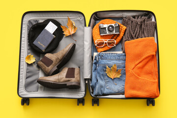 Open packed suitcase with clothes, accessories, shoes and autumn leaves on yellow background