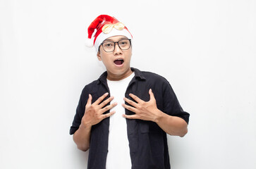 happily surprised young asian man wearing santa claus hat and christmas glasses celebrating christmas. young asian man in santa claus suits making wow gesture. christmas and holiday concept.