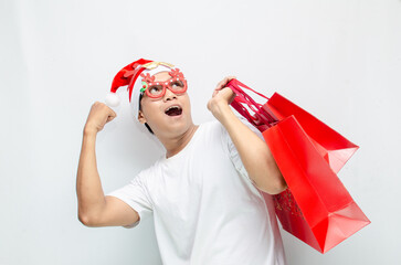 euphoric young asian man wearing santa claus hat and christmas glasses carrying red shopping bag...