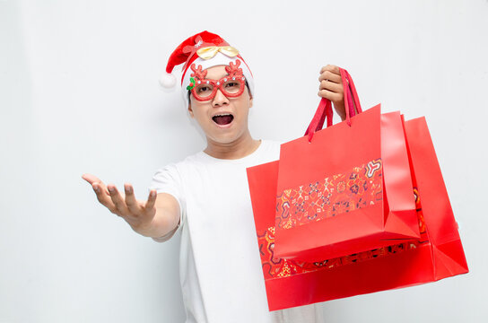 euphoric young asian man wearing santa claus hat and christmas glasses carrying red shopping bag and making yes gesture. christmas sale.