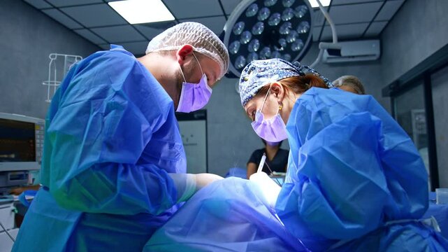 Female surgeon and assisting male medic stand above the patient. Doctor applies electric surgical tool device performing operation on thyroid.