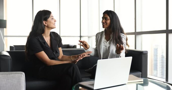 Two cheerful diverse female office friends talking on work break in office lobby, co-working cafe at laptop, discussing fashion, clothes, meeting for offline communication, brainstorming, teamwork