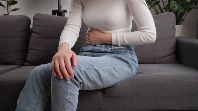 Flatulence ulcer young woman sitting on couch holding on belly, stomach pain from food poisoning, abdominal pain and digestive problem, gastritis or diarrhea. Abdomen inflammation, menstrual period