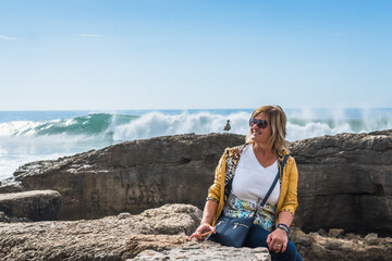 A middle-aged woman sits on the shore of the Atlantic Ocean, watching the waves crash against the shore and create a white splash. Blue pants, yellow jacket and sunglasses. Blue sky.