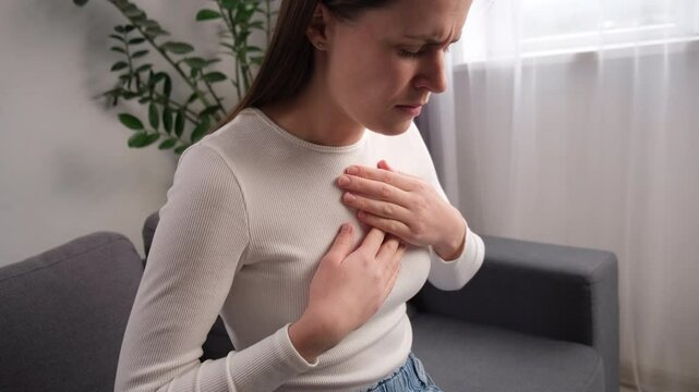 Young woman sit on couch have symptom gastroesophageal, esophageal, stomach ache and heartburn pain hand on chest from digestion problem after eat food. Acid reflux disease. Healthcare medical concept