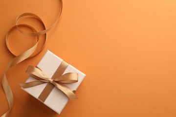 Beautiful gift box with golden bow and ribbon on orange background, top view. Space for text
