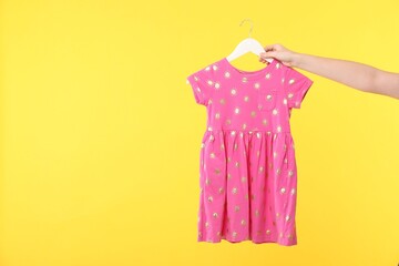 Woman holding hanger with pink baby dress on yellow background, closeup. Space for text