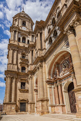 Fragments of majestic facade of Malaga Cathedral (1782), Baroque and Renaissance architecture with grand columns, detailed carvings, and archways under a clear blue sky. Malaga, Andalusia, Spain.