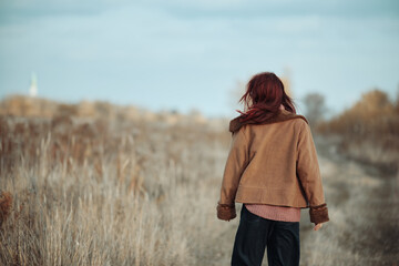 Teenage girl with red hair walking away in a rural field, wearing a brown coat and pink sweater, on an autumn day.