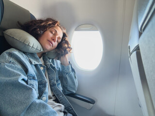 Rest on plane. Young woman napping sitting by porthole in business jet using neck pillow for...