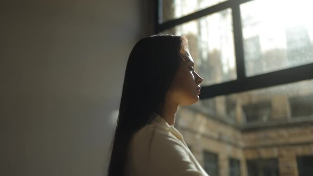 A beautiful girl by the window raises her head to a ray of light in a dark room, study, work, hope, melancholy, loneliness, confidence, a new stage