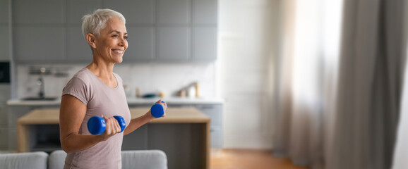 A smiling older woman focuses on her workout, lifting blue dumbbells in a well-lit kitchen. The cozy environment reflects her commitment to fitness and health.