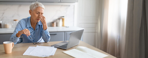 A senior woman sits at a kitchen table, rubbing her temples in frustration while looking at her...