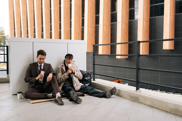Remote view of successful kind businessman and elderly homeless man finding common ground as sharing simple meal sitting together on urban street. Concept of social issues and economic disparity.
