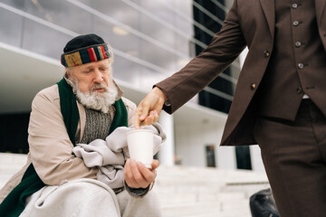 Closeup of sadness elderly homeless man sitting on urban steps and receiving alms in paper cup from unrecognizable businessman in suit, illustrating social disparities and acts of kindness.