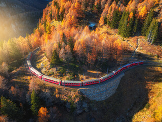 Aerial view of red train traveling through swiss alps mountain pass in fall. Scenic railway winds...