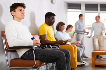 Casual young guy sitting at hall of civil service building, waiting to speak with representative of...