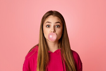 Teenage girl with wide open eyes. Blowing bubble of chewing gum, on pink background, studio shot, free space