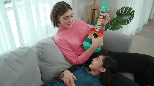 Asian cute daughter listening acoustic music while lie on her mother lap at sofa. Caucasian mother playing colorful ukulele while cute child looking at her parent. Family recreation concept. Pedagogy.