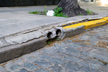 A cracked sidewalk featuring a yellow curb with a tree positioned in the background providing a...