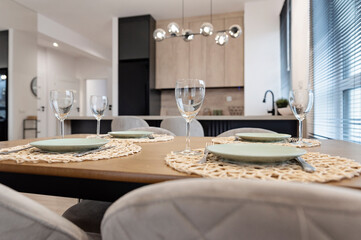 Modern dining room interior with kitchen with wooden counter and dining table with empty plate and glass. Design furniture with stylish glass lapm over the table. Real estate.	