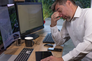 un homme de 50 ans est en télétravail chez lui dans son bureau devant son ordinateur. Il travaille beaucoup et il est fatigué. Il se prend la tête dans les mains.	