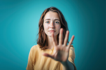 Fototapeta premium Young woman with a serious expression raises her hand to stop workplace harassment, advocating for a respectful and safe work environment. Blue background. Awareness poster.