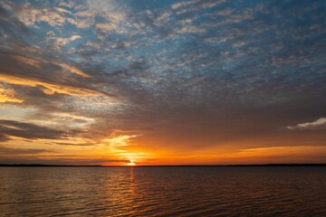 Fiery orange and yellow clouds filling the sky over the calm water of a lake as the sun sets on the distant horizon.