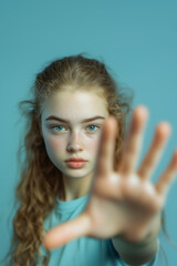 A young girl with fair skin, freckles, and wavy hair gazes intently at the camera, her hand raised in a stop gesture to protest against bullying. Light blue shirt. Soft teal background. Awareness post