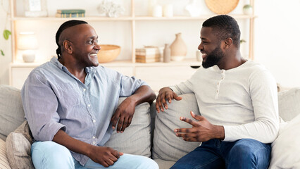 Obraz premium Adult Son And His Black Senior Father Talking While Relaxing On Couch At Home, Smiling To Each Other, Free Space