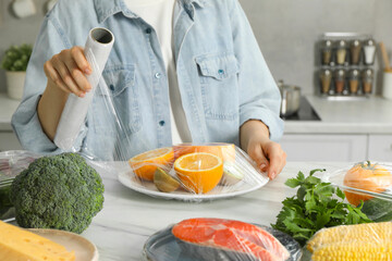 Woman putting plastic food wrap over plate with fresh fruits at white table in kitchen, closeup