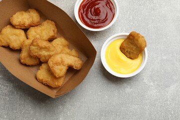 Delicious chicken nuggets in carton box and sauces on gray textured table, flat lay
